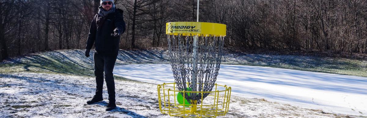 A person throwing a disc into a disc golf net, with snow on the ground.