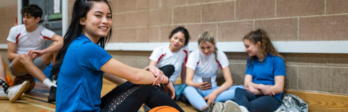 A shot of a female teenage athlete sitting on the floor during half time of a basketball game. She is smiling and looking into the camera. She is socializing with her team mates with a basketball by her side.
