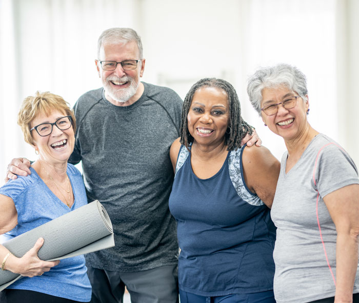 Four senior adults stand in a row smiling at the camera in workout clothing. A woman also holds a rolled up yoga mat.
