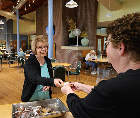 Two senior women smile at each other while one hands the other a wrapped cookie in a seniors centre
