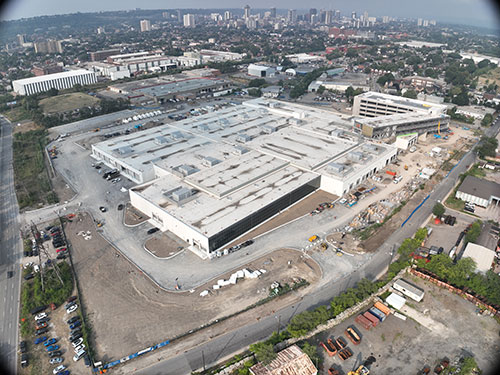 Photo taken August 5, 2025, looking southwest towards downtown Hamilton. Driveway and drive aisle curbing continue. Minor grading commenced. Windows are being installed on the elevated Administration building located near the parking structure on the right side of the picture.