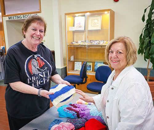 Two older women smiling at the camera, one is shopping for the knit wear the other woman has displayed at her craft table