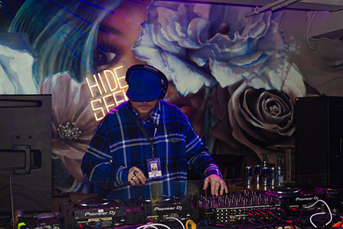 A person djing with lots of colourful artwork and a neon sign behind them