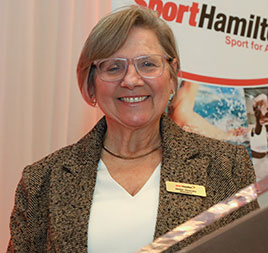 Photo of Helen Downey: A woman with short dark blonde hair and glasses, wearing a blazer and standing behind a podium, smiling at the camera.