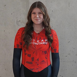 Portrait of Leighton Underhill: Young woman in a red and black cycling outfit, with long brown hair, smiling at the camera.