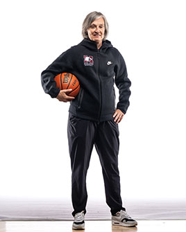 Portrait of Theresa Burns - a woman with chin length grey hair, wearing a McMaster basketball sweatsuit and holding a basketball under her arm, smiling at the camera.