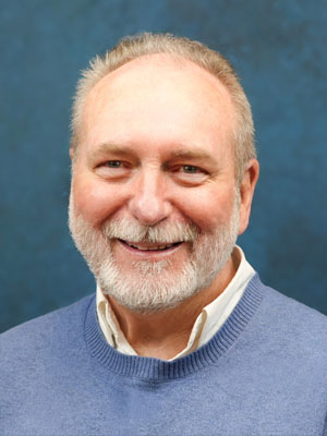 Smiling man with light grey hair and beard, wearing a white collared shirt under a light blue sweater
