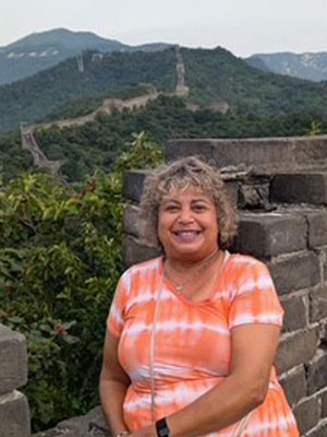 Smiling woman with grey hair, leaning against a brick half-wall overlooking a mountain range and trees