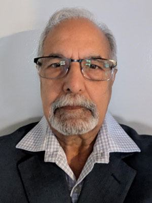 Man with brown skin, glasses and white/grey hair and a beard wearing a collared white shirt and a black blazer