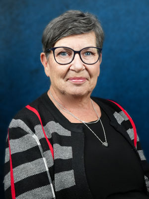 A woman wearing glasses with short dark grey hair, wearing a black shirt and a black and grey striped cardigan with pink lines on it