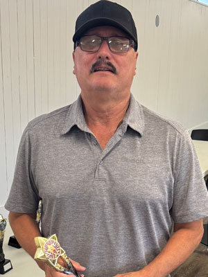 Man with dark mustache wearing a grey collared shirt and a black baseball cap, holding a brass trophy