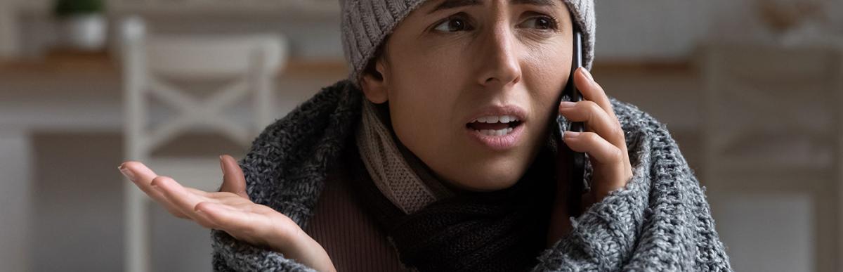 Young woman wear a knit hat and under a blanket, calling landlord to complain about low temperature