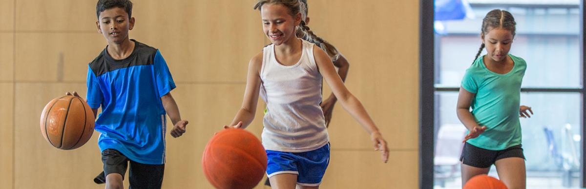 A multi-ethnic group of elementary age children are dribbling basketballs down the court in the gym.