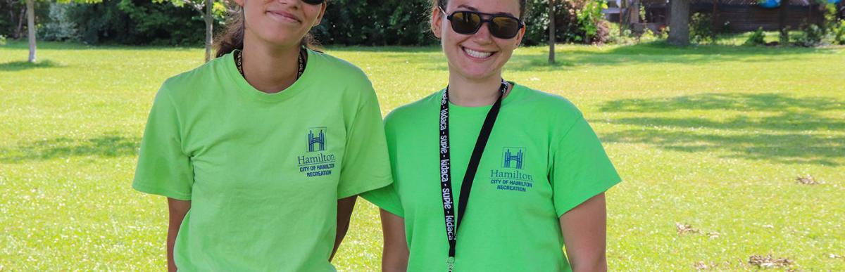 Two people in green Hamilton recreation t-shirts and sunglasses, standing in a park
