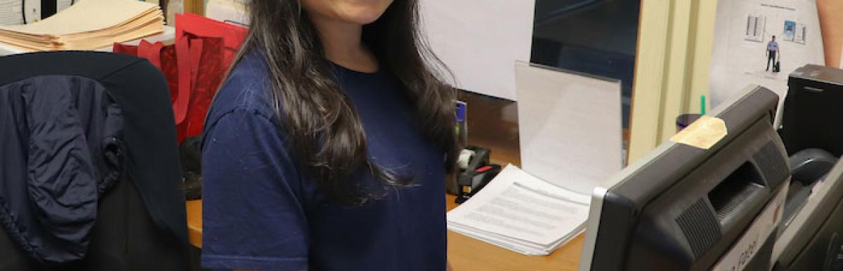 Young woman standing at a desk with a computer on it, and smiling at the camera