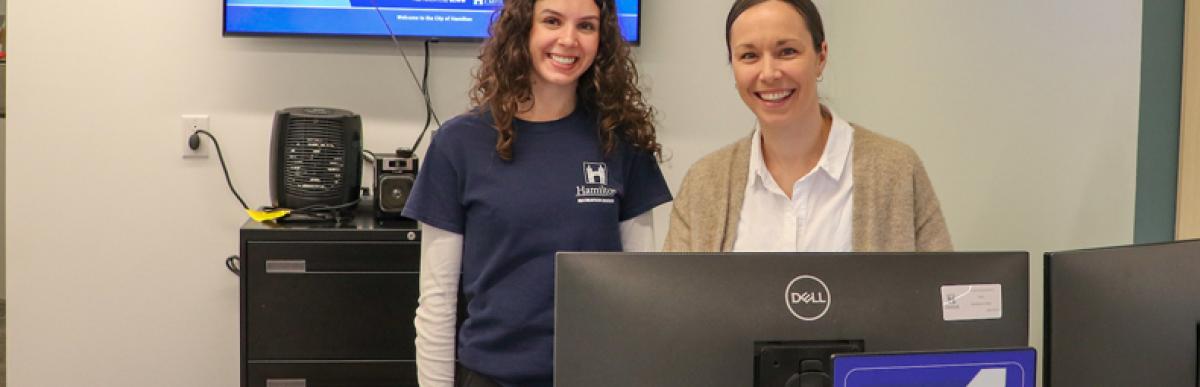Two recreation staffers standing behind a computer monitor and a desk.