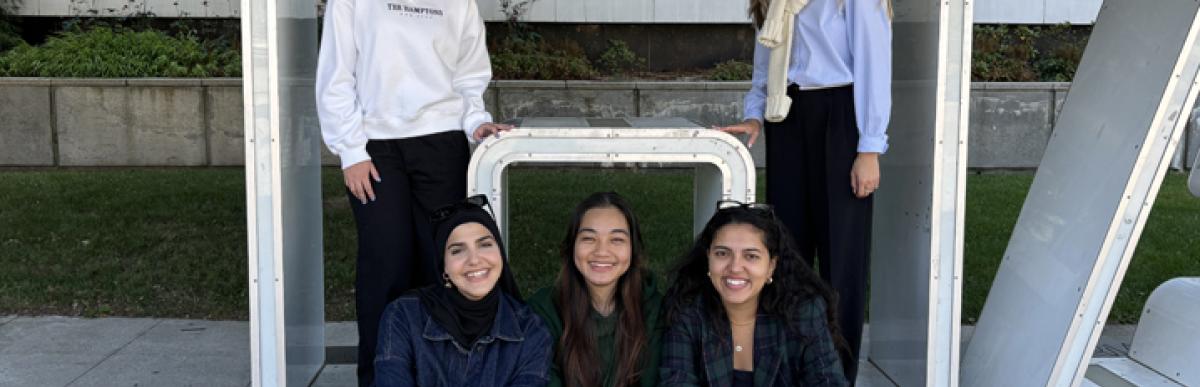 Five young people smiling and standing in front of the H in the Hamilton signature sign