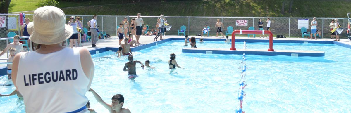 Lifeguard with back to camera watching over many kids and adults swimming in an outdoor pool