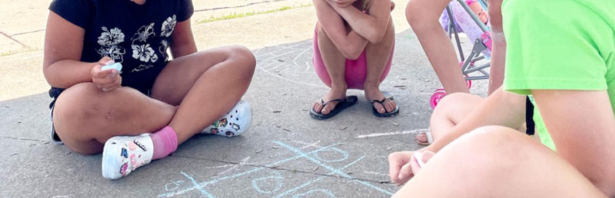 A teenage Supie sits on the ground at a park with several children, drawing with chalk on the concrete.