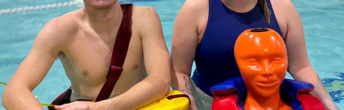 Two teenage lifeguards in an indoor pool with a lifesaving dummy and floating board