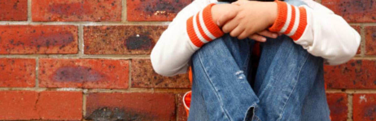A child sitting on the ground with knees up and arms on knees, back against a brick wall, hiding their face in their arms.