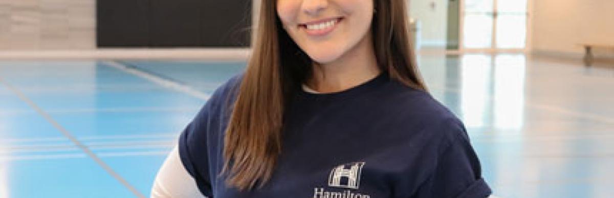 Young woman in a Hamilton Rec Staff tshirt smiling at the camera, inside a Recreation centre gym
